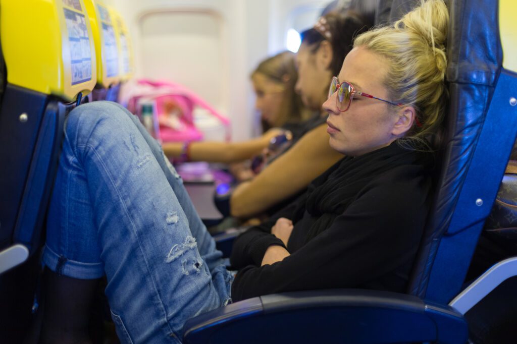 An uncomfortable blonde lady in an aisle seat is trying to sleep and showing the importance of Long Haul Flight Tips and why seat choice matters.