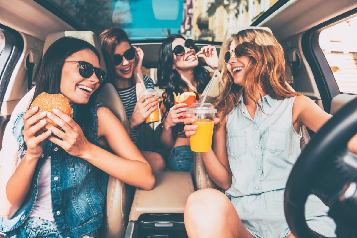 Enjoying their lunch in the car. Four beautiful young cheerful women looking at each other with smile and eating road trip snacks while sitting in car