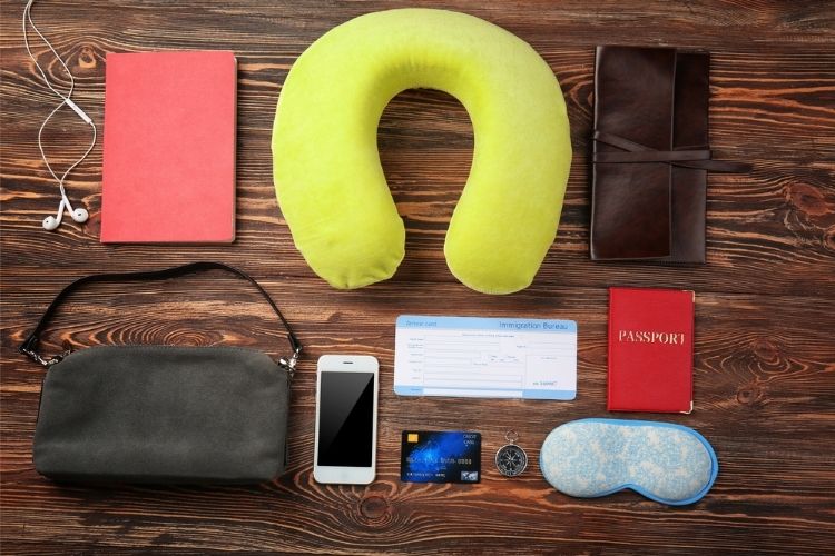on table: travel pillow, wallet, passport, phone, sleep mask, pure, compass, headphones and note book